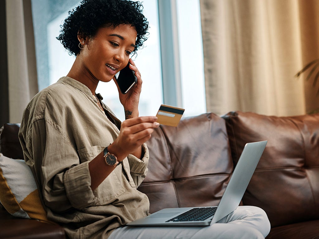 Shot of a young woman looking at her credit card while talking on the phone and sitting with her laptop