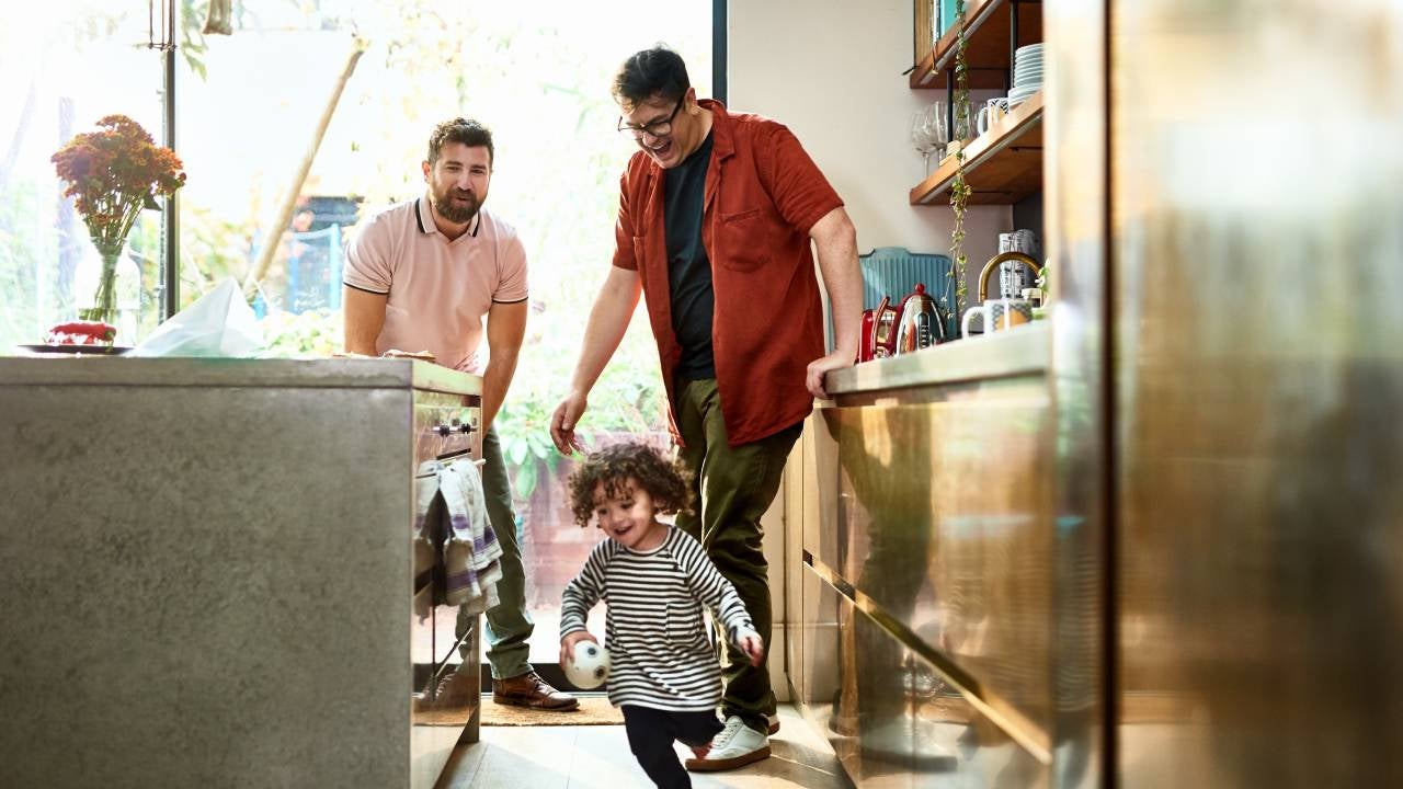 Two fathers watching their toddler playing with ball in kitchen